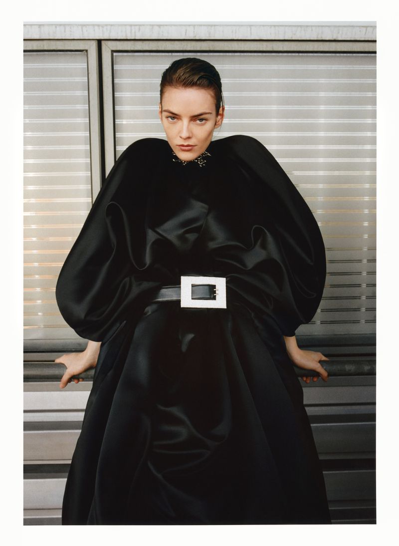 Fashion shot of a female model in a black dress with a large white belt buckle, posing against a window with horizontal blinds.