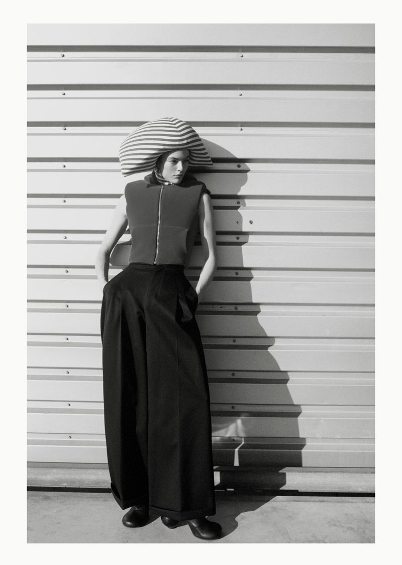 An adult female model poses in an editorial fashion shot wearing a large striped hat, vest, and wide black pants against a corrugated wall.