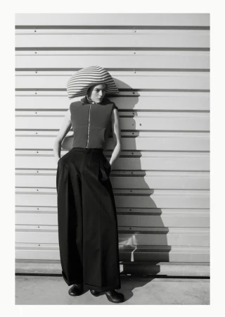 An adult female model poses in an editorial fashion shot wearing a large striped hat, vest, and wide black pants against a corrugated wall.