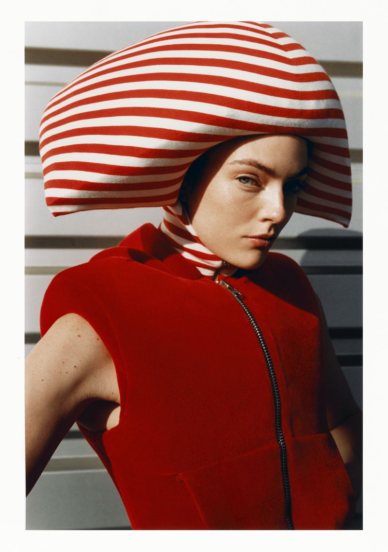 Striking portrait of a woman with a red and white striped hat, wearing a red sleeveless top, against a neutral background with horizontal lines.