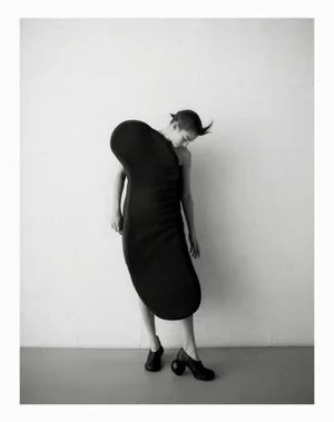 Model wearing a sculptural black dress with rounded volumes and black shoes standing against a plain white wall