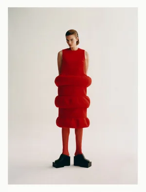Model wearing a structured red dress with horizontal rings and chunky black platform shoes against a white background.