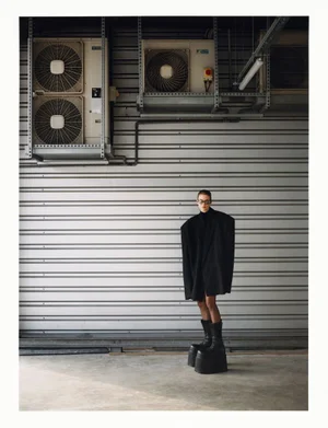 Model wearing oversized black coat and platform boots standing before industrial air conditioning units