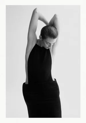 Model with slicked-back hair wearing a dark sleeveless top and skirt, arms raised overhead against a plain white background.