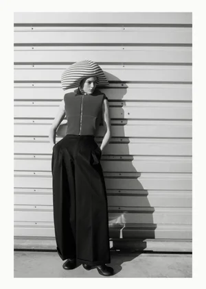 Model wearing a striped sculptural hat and high-waisted trousers leaning against a corrugated metal wall in a black and white photograph.