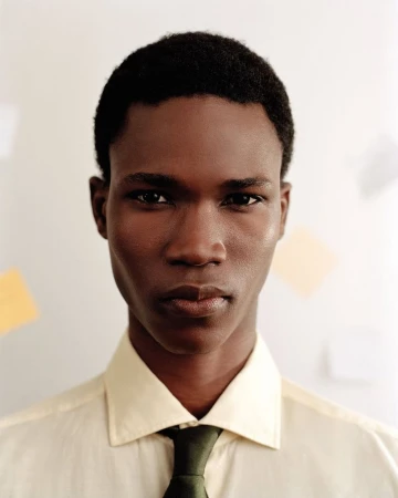 Portrait of a serious African American adult male wearing a yellow shirt and dark tie against a white wall with blurred papers.
