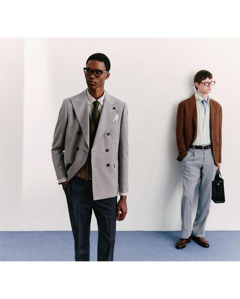 Two men in suits pose against a white wall. One man wears a grey suit, the other a brown jacket, classic eyewear, and a briefcase.