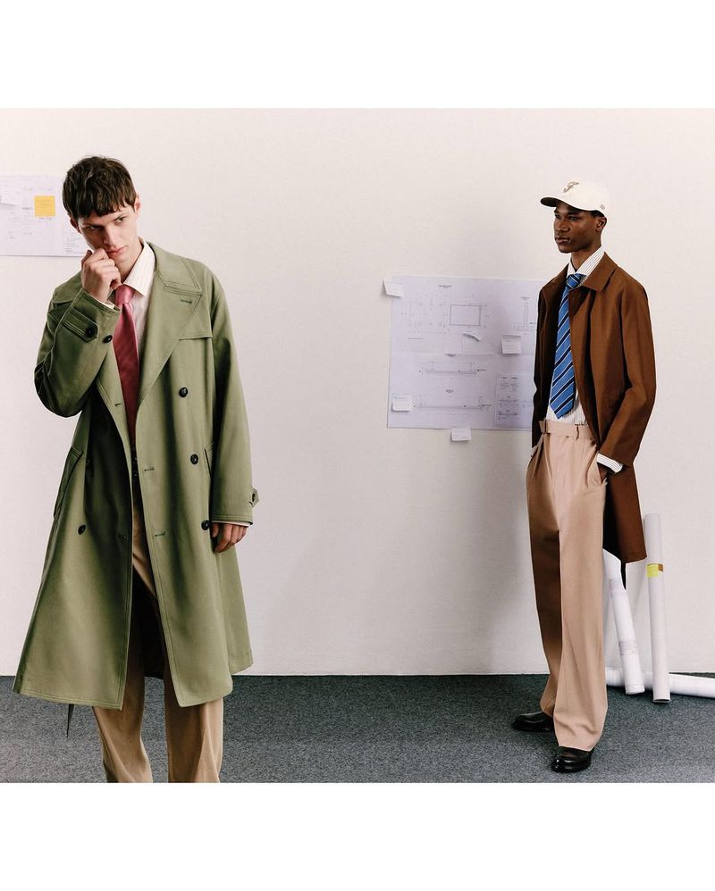 Two men in fashionable trench coats and dress pants stand in front of a white wall with architectural drawing and blueprints.