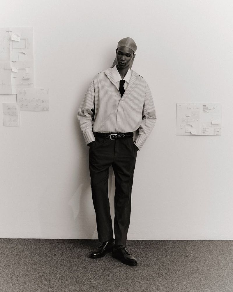 Portrait of Black male model wearing striped shirt, tie, and head covering, standing in a minimally decorated studio. Fashion editorial.