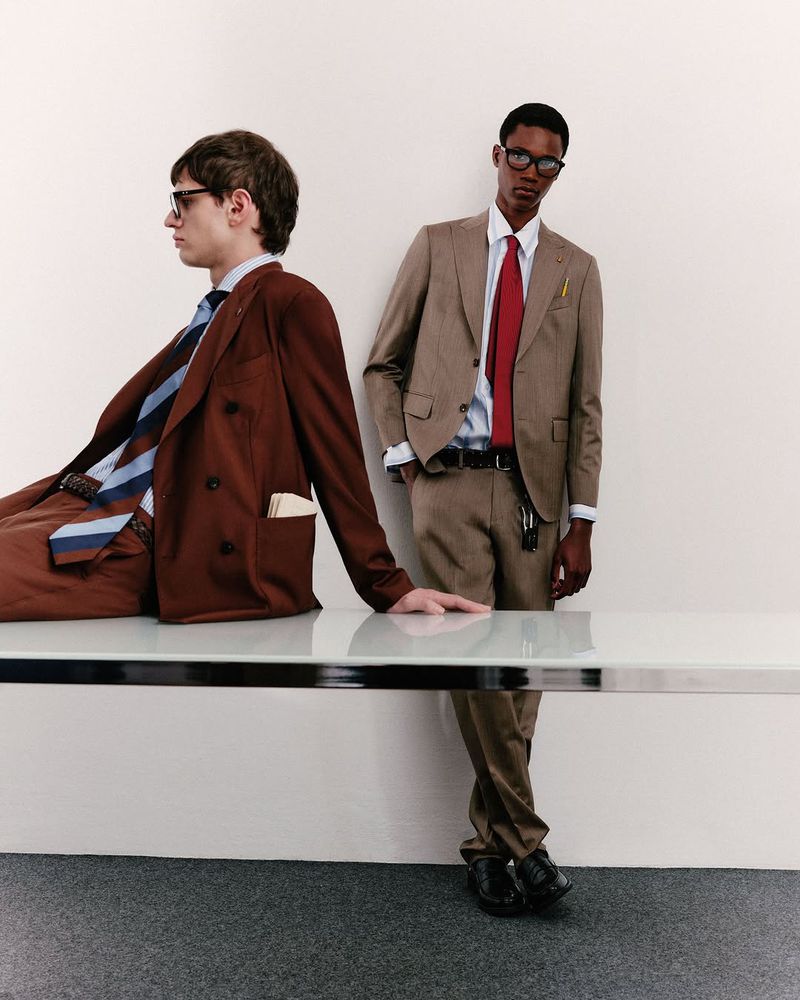 Two stylish men in suits pose indoors. One man sits on a glass table while the other stands, leaning against the wall in a studio shot.