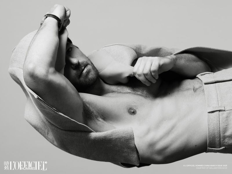 Black and white portrait of a shirtless male model, lying down and facing the camera, wearing trousers, and a hooded jacket draped over the body.