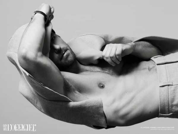 Black and white portrait of a shirtless male model, lying down and facing the camera, wearing trousers, and a hooded jacket draped over the body.
