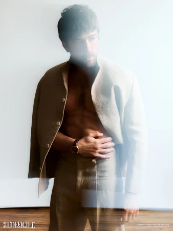 A light-skinned male model poses shirtless in a khaki suit jacket and pants against a bright white background in a studio fashion shot.