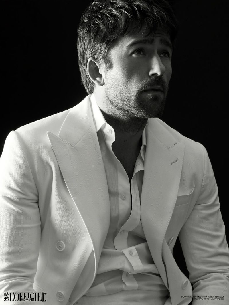 Monochrome portrait of adult male with facial hair wearing a white suit and shirt against a black backdrop.