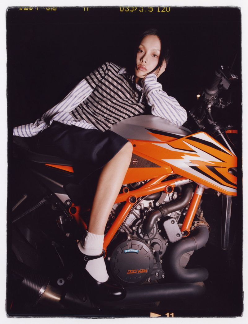An adult female poses with an orange and black motorcycle indoors, showcasing a blend of fashion and automotive elements in a studio setting.
