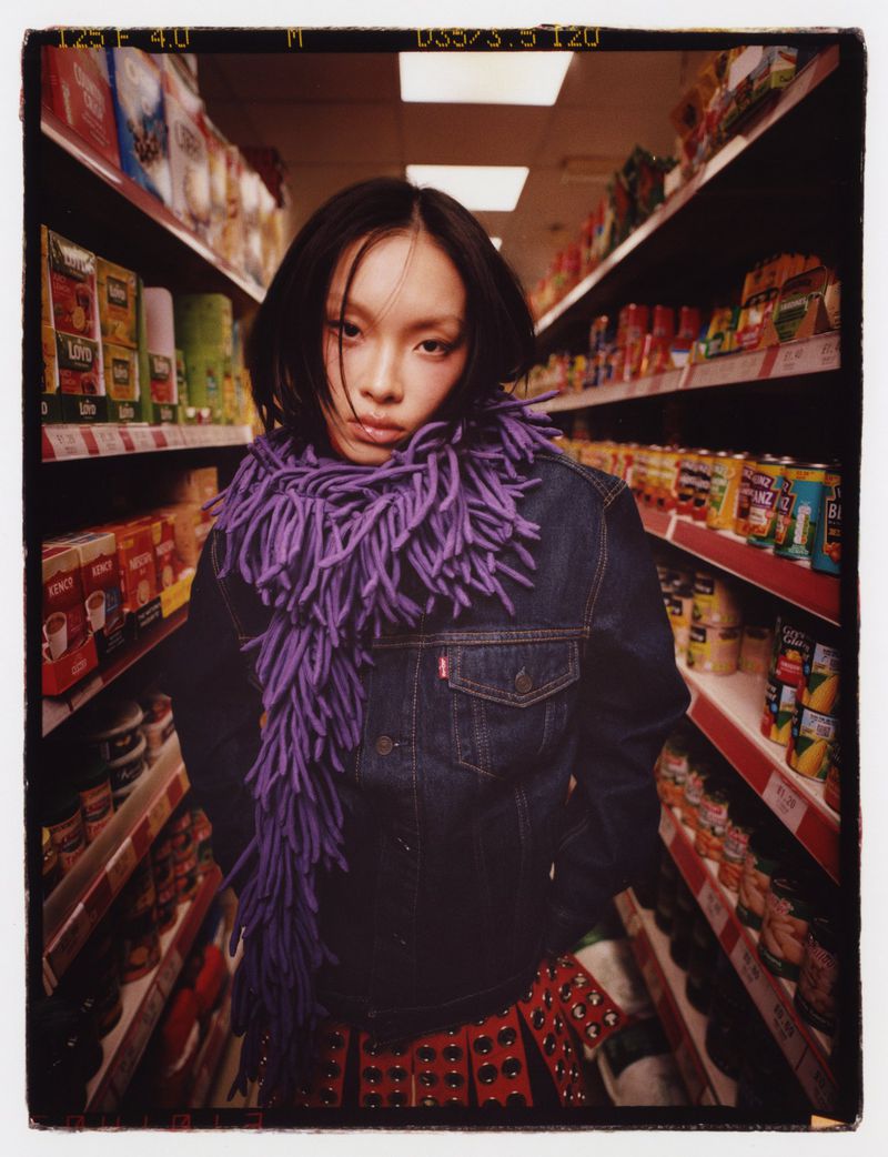 Young Asian woman in denim jacket and purple fringe scarf stands in a grocery store aisle lined with shelves of food items.