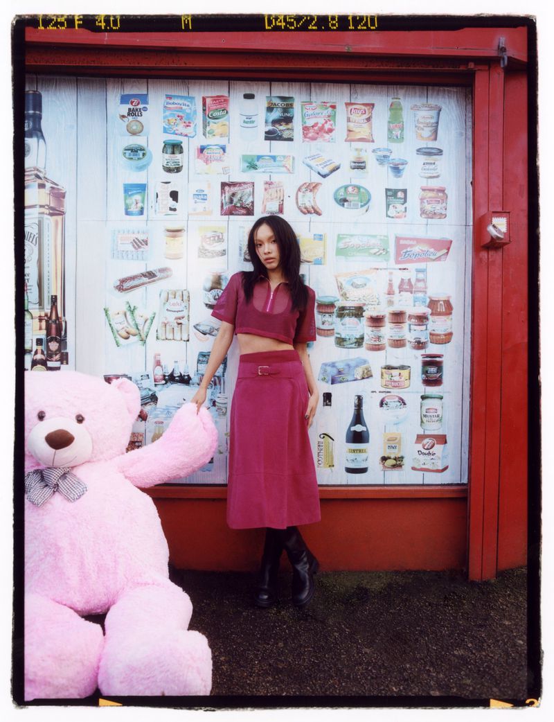 An adult woman poses with a large pink teddy bear in front of an outdoor wall covered in vintage-style food product images.