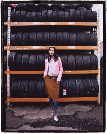 Fashionable woman posing in front of wall of tires on shelving unit in outdoor setting wearing a skirt, jacket, and knee-high socks.