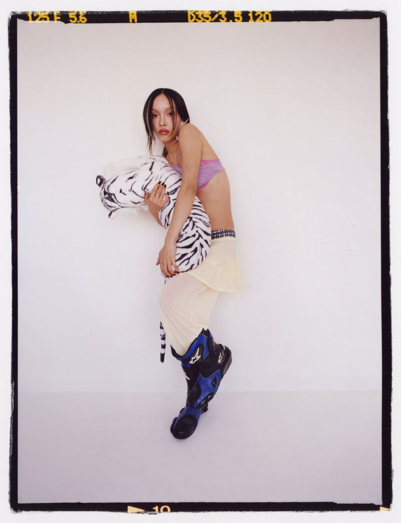A slender young woman in casualwear strikes a candid pose in a studio, cradling a stuffed tiger, and looking toward the camera.