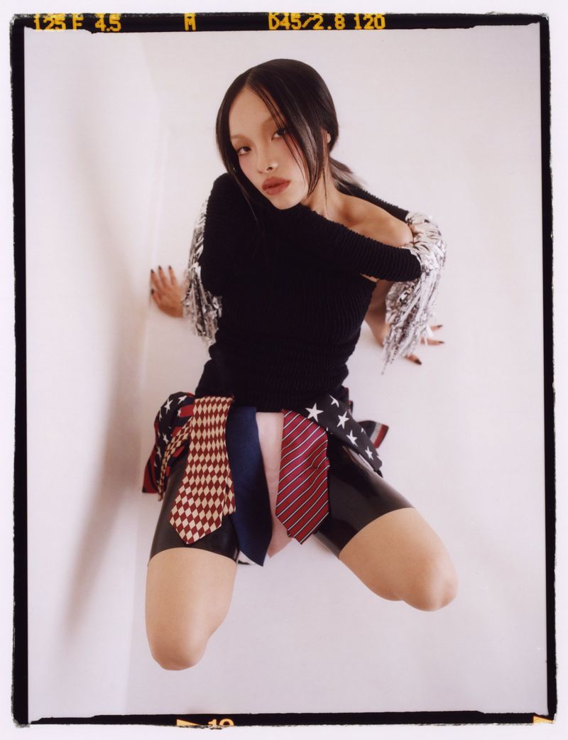 Woman in a black top and patterned skirt posing in front of a white wall. The skirt is made of ties with different patterns.