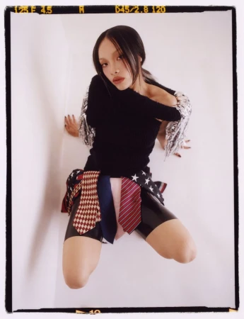 Woman in a black top and patterned skirt posing in front of a white wall. The skirt is made of ties with different patterns.