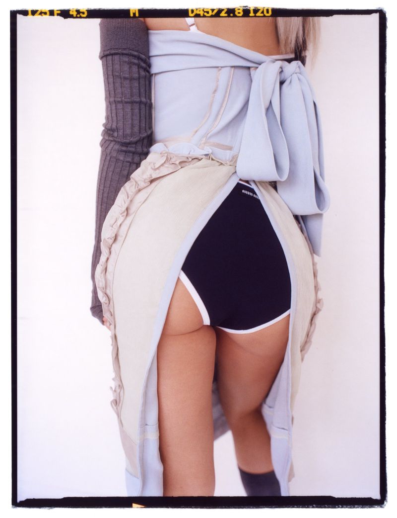 From the back, woman poses in a draped light blue and beige garment with a black and white athletic shorts underneath on white.
