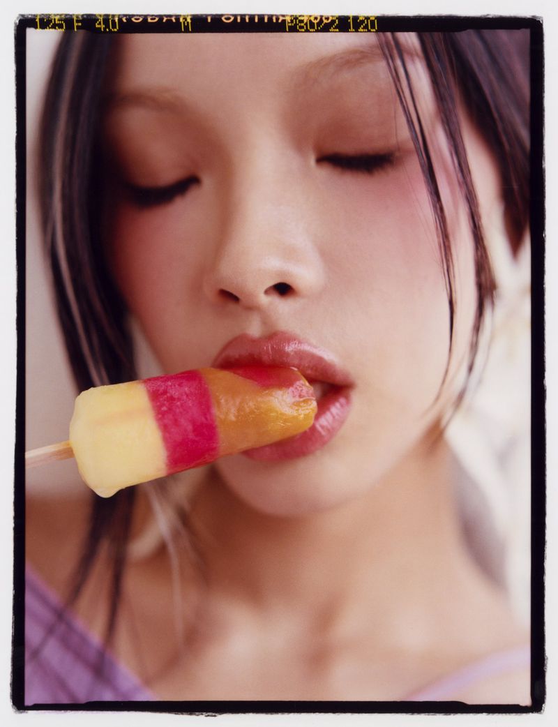Close-up of an adult female with fair skin and dark hair biting into a tri-color ice pop against a simple light studio backdrop.