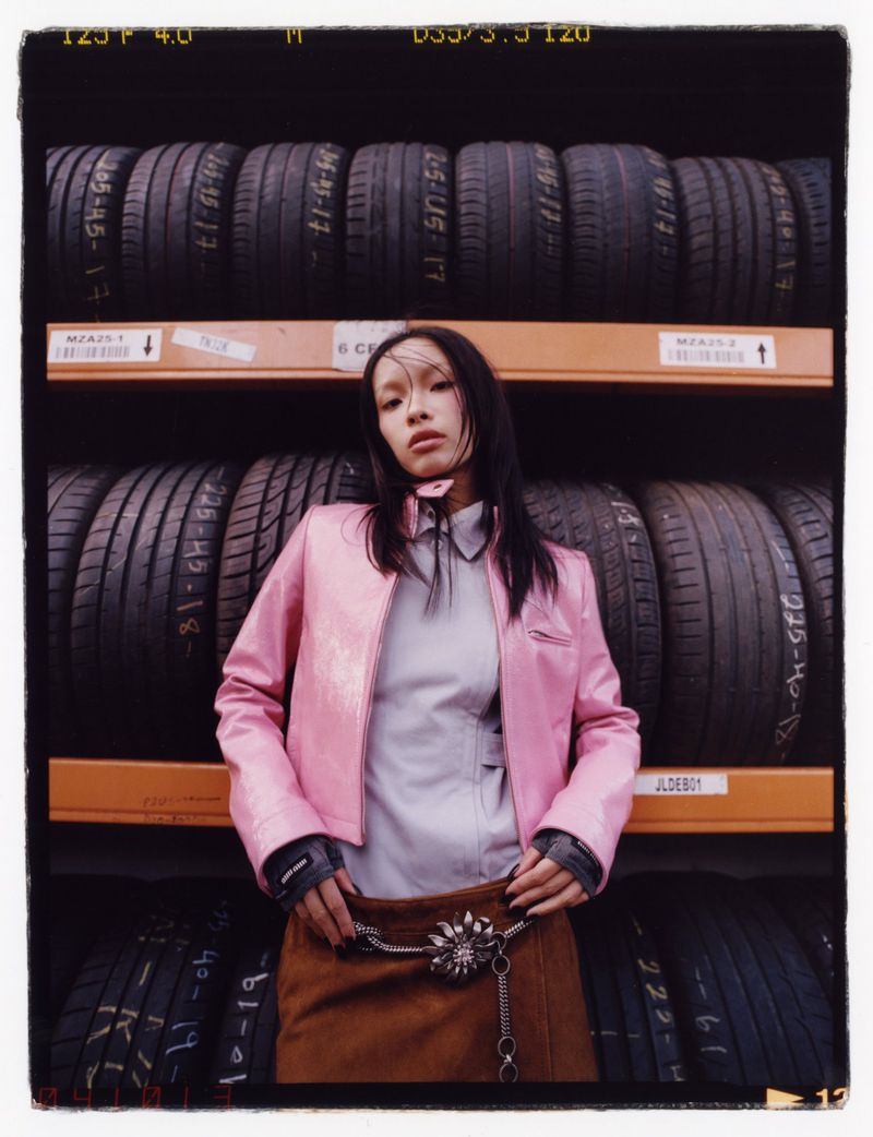 Young woman with dark hair poses indoors wearing a pink jacket and brown skirt, standing in front of stacked tires on shelves.
