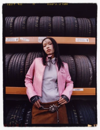 Young woman with dark hair poses indoors wearing a pink jacket and brown skirt, standing in front of stacked tires on shelves.