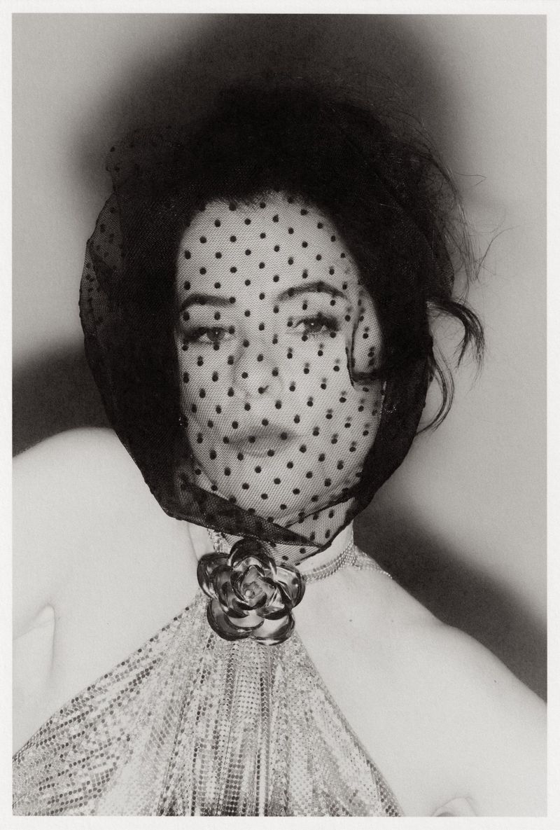Black and white portrait of an adult woman with dark hair, a dotted veil, rose brooch, and textured dress against a diffused background.