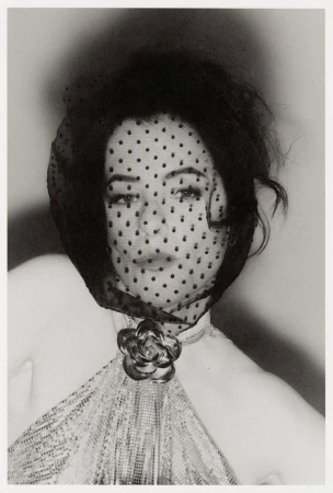Black and white portrait of an adult woman with dark hair, a dotted veil, rose brooch, and textured dress against a diffused background.