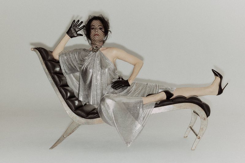 Elegant woman in a silver gown and black veil poses in a modern chair against a white backdrop, showcasing fashion and style.