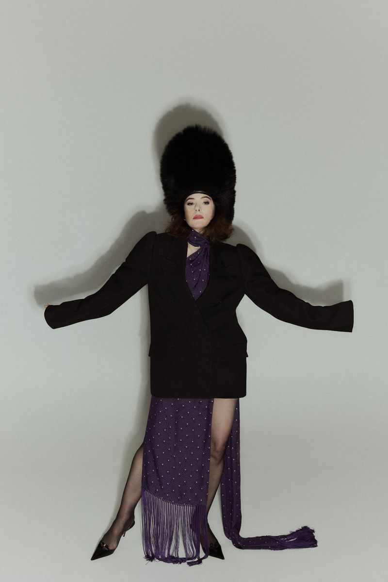 A woman stands in a light grey studio wearing a black blazer, purple dress, black stockings and heels, and a tall black military hat.