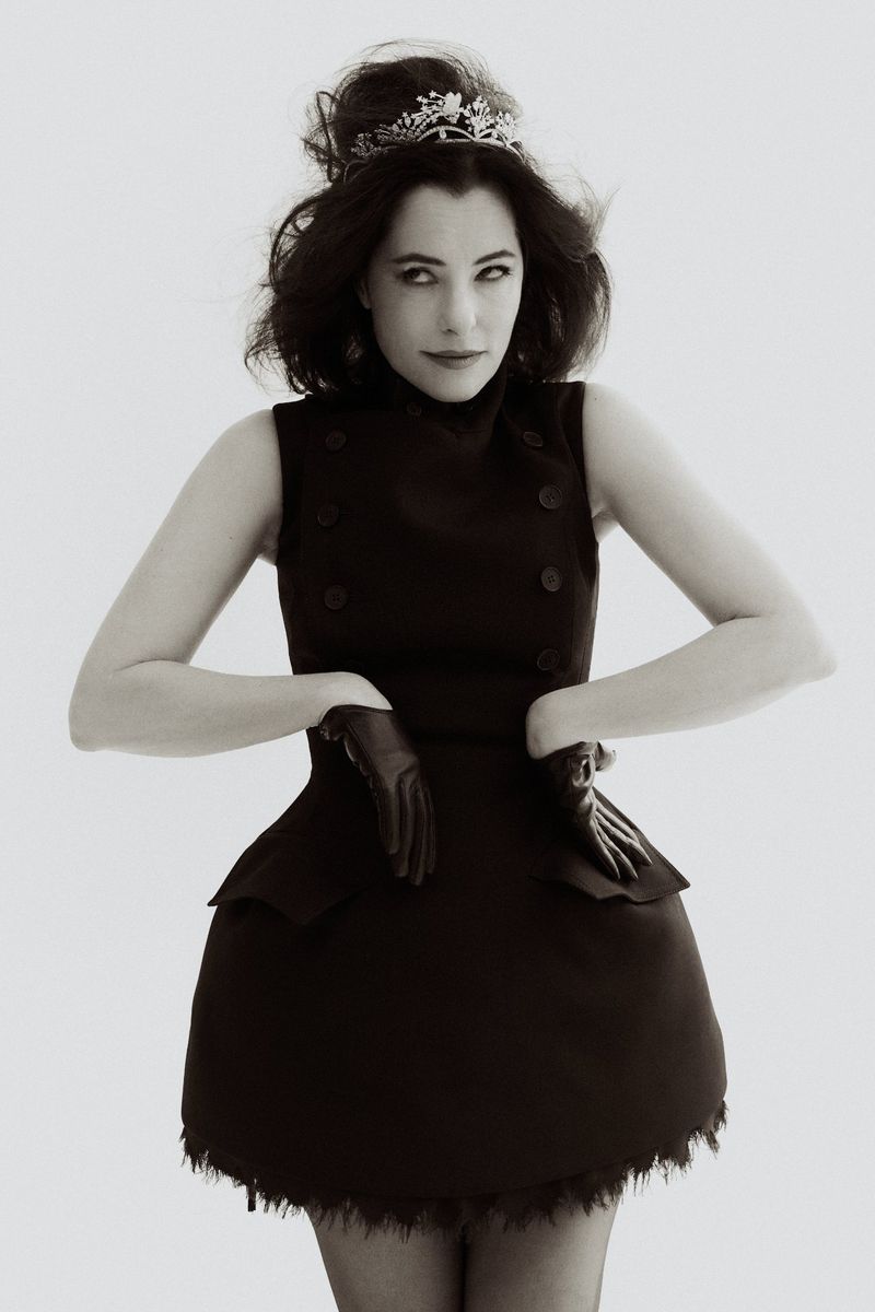 Parker Posey wearing a dark dress with leather gloves and a tiara against a white seamless background