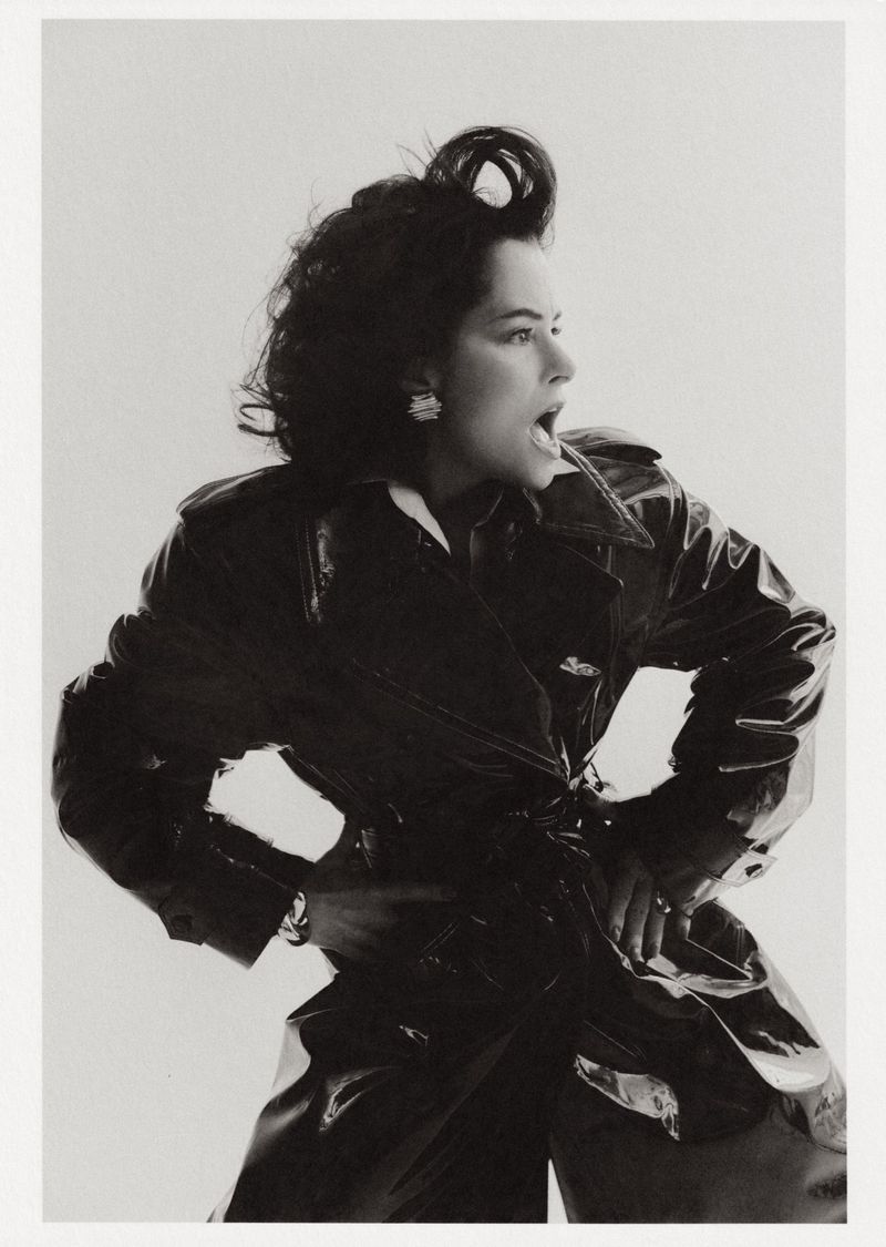 Black and white portrait of Parker Posey wearing a glossy trench coat with hands on hips against a plain background for WWD.