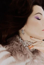 Close-up portrait of Parker Posey wearing a sparkling necklace, crystal earrings, and a textured coat against a dark background.