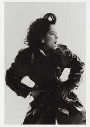 Black and white portrait of Parker Posey wearing a glossy trench coat with hands on hips against a plain background for WWD.