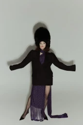 Parker Posey in a black blazer, purple patterned skirt, and large faux fur hat against a white seamless backdrop.