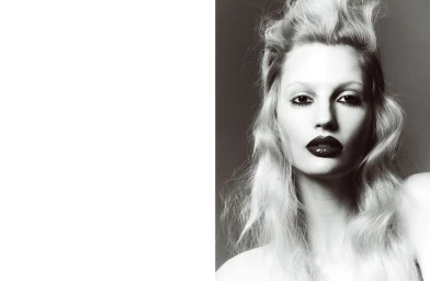Close-up portrait of a model with voluminous light-coloured hair, dark heavy eye makeup, and glossy dark lips against a neutral background.