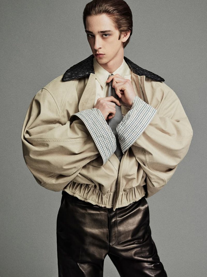 Portrait of a young adult male tying his tie and wearing an oversized jacket and leather pants against a gray studio backdrop.
