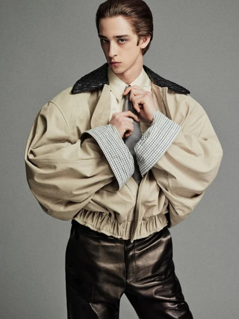 Portrait of a young adult male tying his tie and wearing an oversized jacket and leather pants against a gray studio backdrop.