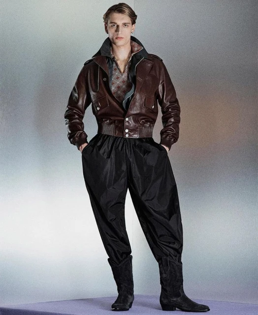 Fashionable adult male model in a brown bomber jacket, black parachute pants and boots posing in a photo studio with neutral lighting.
