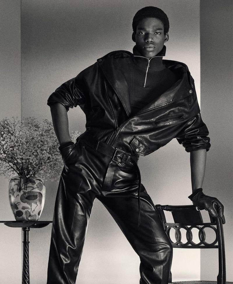 Black and white image of model in leather jacket and pants leaning on chair with vase of flowers in the background. Fashion photography.
