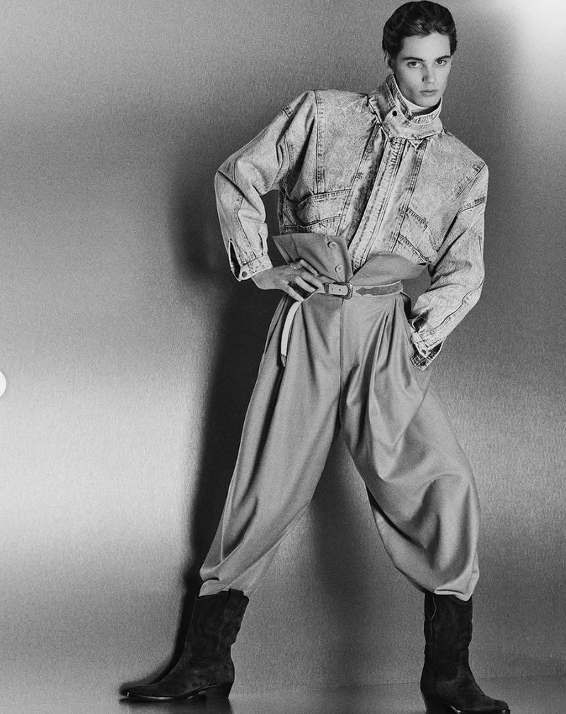 Fashion model poses in light blue denim jacket, high-waisted trousers, and black boots against a gradient gray backdrop in a studio setting.