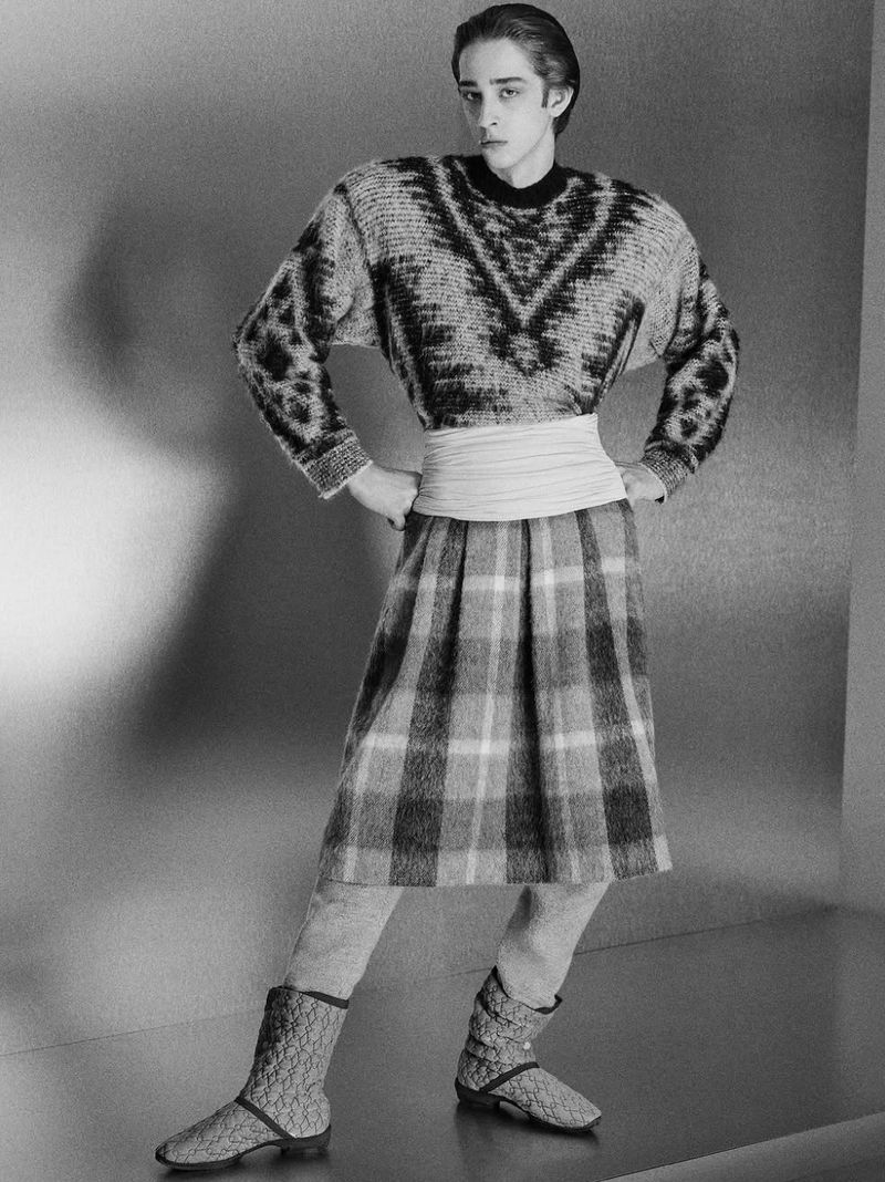 A male fashion model poses in a gray, patterned sweater and plaid skirt in a studio against a grey background, hands on hips.