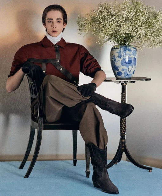Stylish young adult male poses in a maroon button-down shirt and brown pants, seated in an elegant chair near a vase of flowers.