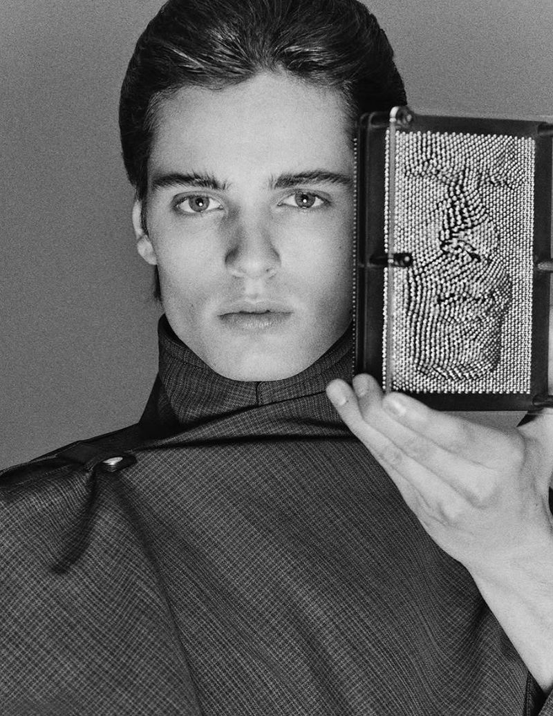 Portrait of a young man holding a pin impression toy showing the impression of a face in black and white with a grey backdrop.