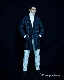 Man wearing a dark double-breasted patterned coat, white turtleneck, and light trousers standing against a black background.