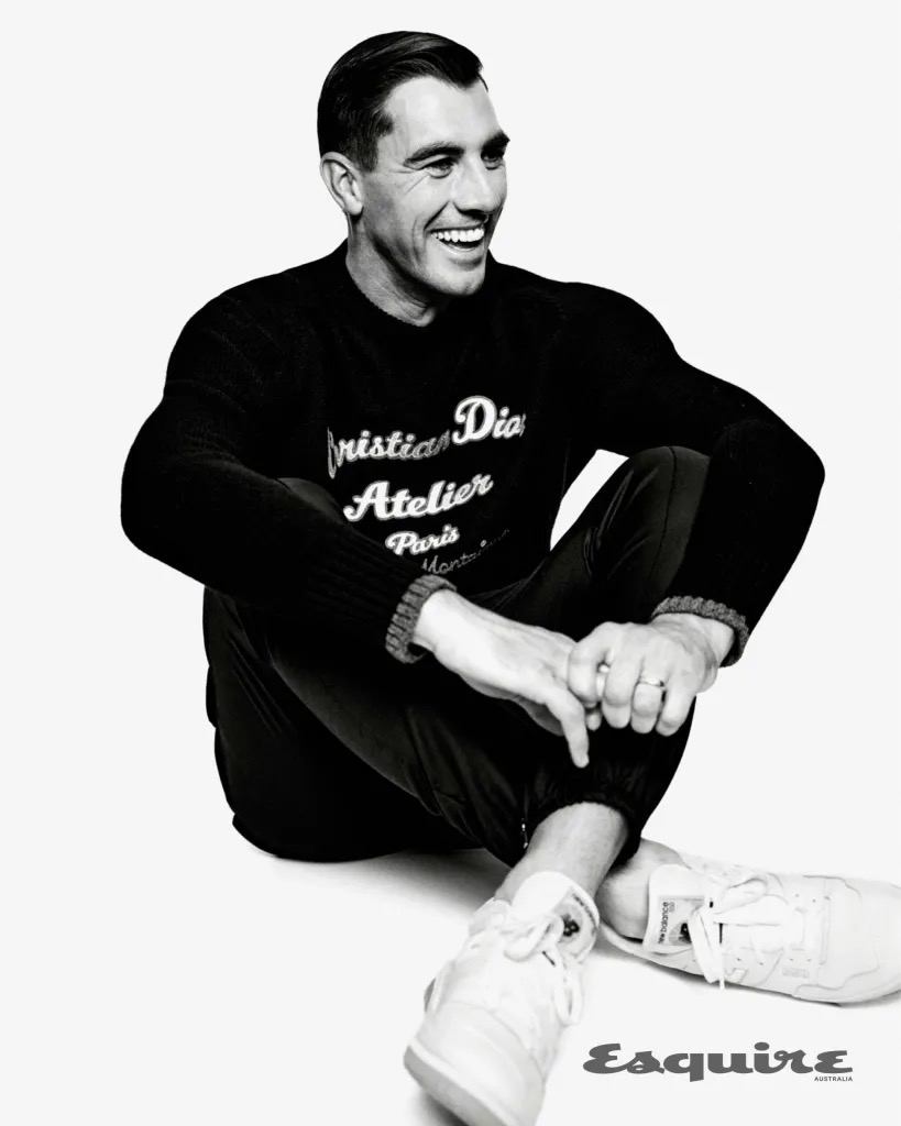 Man seated on a white background wearing a black sweater with white lettering and white sneakers