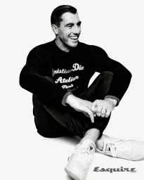 Man seated on a white background wearing a black sweater with white lettering and white sneakers